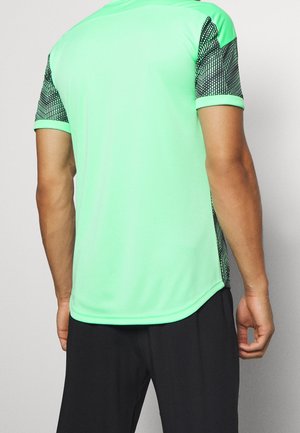 Homme portant une chemise à manches courtes vert vif avec des manches noires à motifs et un pantalon noir, vu de dos.