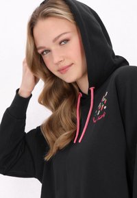 Sudadera negra con cordones rosas, con coloridos acentos bordados. Tejido suave con un corte relajado y un bolsillo tipo canguro.