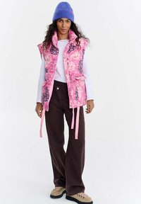 Różowa pikowana kamizelka z kwiatowym wzorem i stójką, noszona na białej bluzce z długim rękawem. Połączona z brązowymi sztruksowymi spodniami i beżowymi sznurowanymi butami.