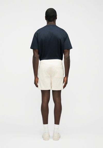 Homme vu de dos portant un t-shirt bleu marine foncé, un short crème avec une poche arrière, des chaussettes blanches et des baskets de couleur claire.