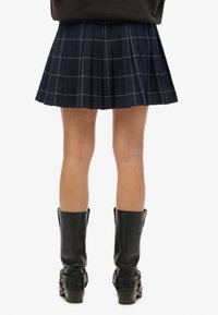 Mini falda plisada azul marino con patrón de cuadrícula blanca, hecha de tela texturizada. Combinada con botas de cuero negras hasta la mitad de la pantorrilla con hebillas.