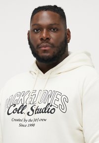 Cremefarbener Hoodie mit großen schwarzen Buchstaben "Jack & Jones Coll. Studio", ausgestattet mit einer Kordelzugkapuze und weichem, strukturiertem Material.
