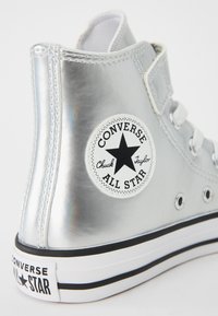 Baskets montantes argentées avec une finition brillante, arborant un patch logo étoile noir, des lacets blancs et une semelle noir et blanc.