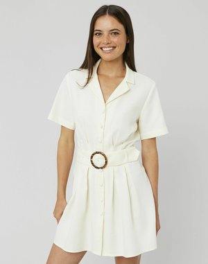 Femme souriante portant une robe blanche à manches courtes, ceinturée avec des boutons et une boucle en écaille de tortue, se tenant contre un fond gris.