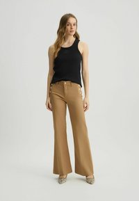 Zwarte geribbelde tanktop, gecombineerd met tan kleurige flared broek met gouden knoopaccenten aan de zijkant. Geribbelde schoenen met slangenprint maken de look compleet.