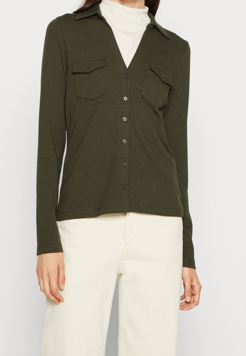Chemise à manches longues vert olive avec un col en V, deux poches poitrines et des boutons noirs. Tissu doux avec une coupe décontractée, associée à un pantalon couleur crème.