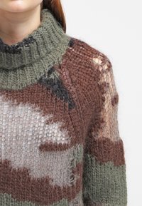 Neulepaita, jossa on paksu vihreä poolokaulus, jossa on ruskean, vihreän ja harmaan sävyinen kamouflagekuvio. Avoimen kudontateknologian rakenne, jossa on läpikuultavia alueita.