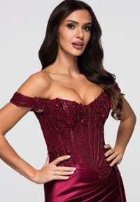Bordowa suknia odkrywająca ramiona z misternymi zdobieniami z koralików i cekinów, dopasowanym gorsetem oraz spływającą satynową spódnicą. Gładka tekstura tkaniny.