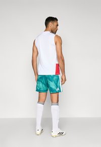 Bela majica brez rokavov z rdečim poudarkom, turkizne kratke hlače z abstraktnim vzorcem, črno-bele črtaste nogavice in beli kopački z rumenimi detajli.