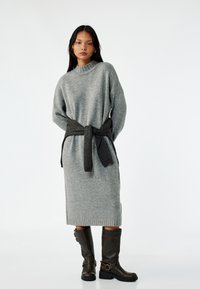 Grijze gebreide jurk met een hoge hals en een donkergrijze riem die om de taille is geknoopt. Het model draagt bruin kniehoge laarzen met gespen.