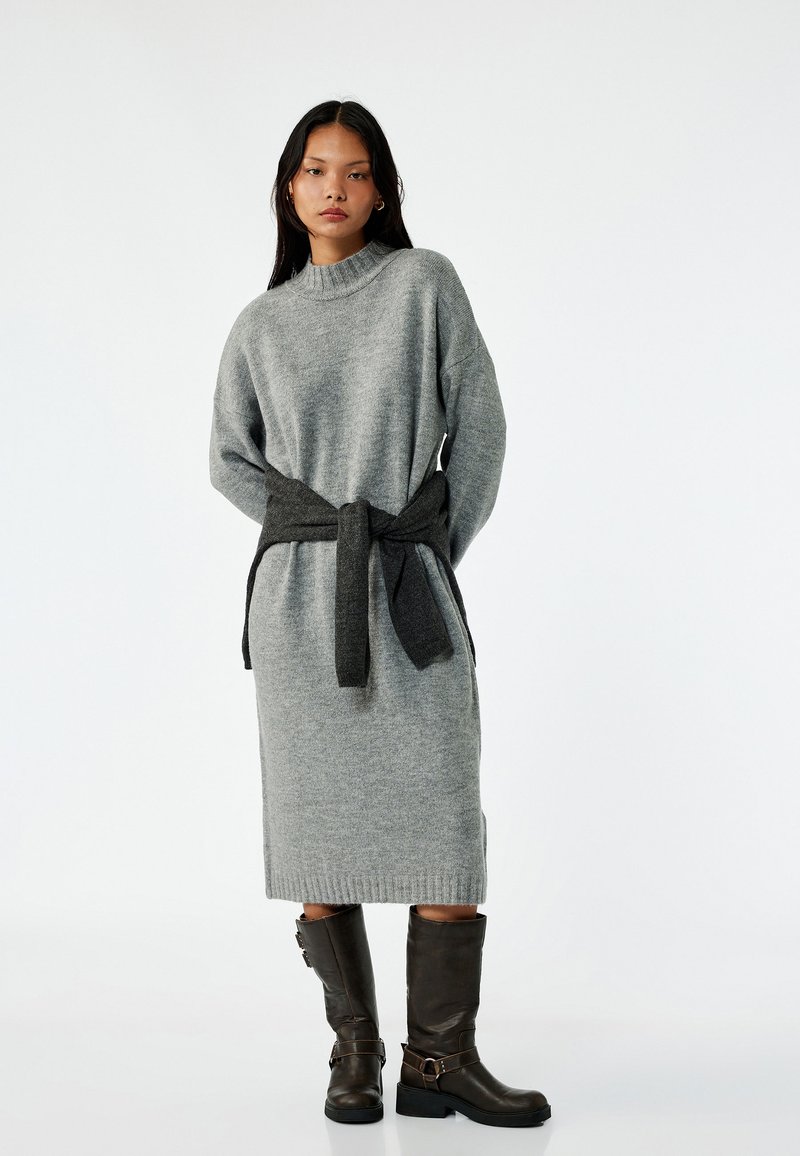 Grijze gebreide jurk met een hoge hals en een donkergrijze riem die om de taille is geknoopt. Het model draagt bruin kniehoge laarzen met gespen.
