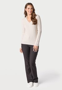 Giovane donna con lunghi capelli castani, vestita con un maglione a trecce color crema, pantaloni neri e sneakers bianche, in piedi e sorridente.