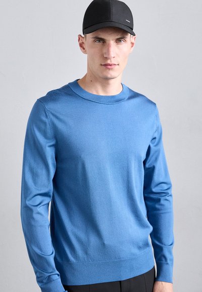 Mėlynas ilgas rankovės megztinis, pagamintas iš glotnaus audinio, su apvalia iškirpte. Modelis dėvi juodą kepurę. Minkšta tekstūra, minimalus dizainas, prigludusi siluetas.