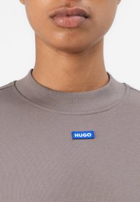 Gray ribbed sweatshirt with a crew neckline, featuring a blue "HUGO" logo patch on the front. Smooth fabric with a fitted design.