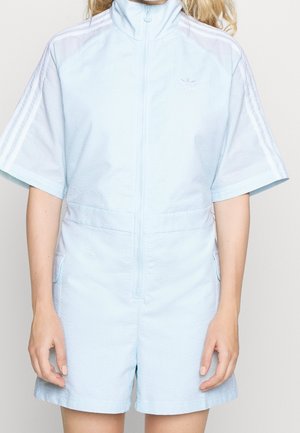 Woman wearing light blue Adidas short-sleeve zip-up jumpsuit with white stripes on sleeves and side pockets, standing against a plain background.