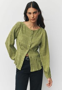 Vihreä teksturoitu pusero, jossa pyöreä pääntie, napitus ja pitkät hihat. Puserossa on poimutettuja yksityiskohtia ja istuva vyötärö. Yhdistä mustien farkkujen kanssa.