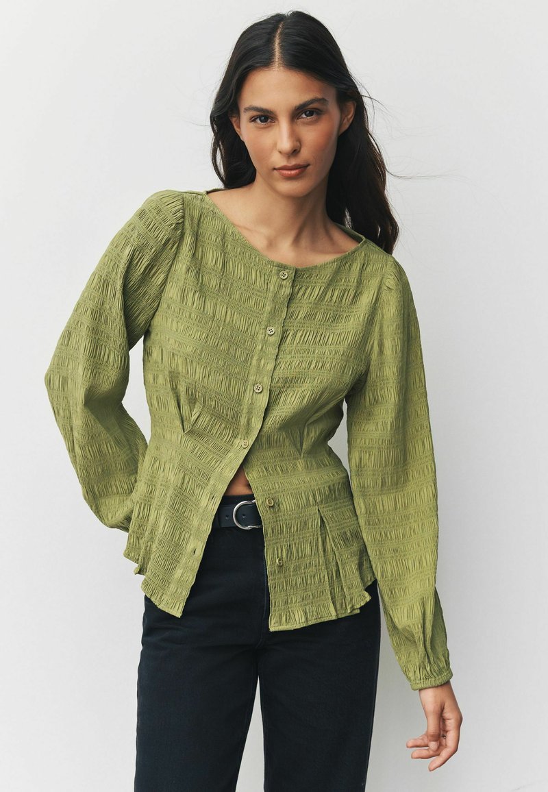 Zielona bluzka o fakturowanej tkaninie z okrągłym dekoltem, zapinana na guziki, z długimi rękawami, ozdobiona marszczeniami i dopasowaną talią. Połącz z czarnymi jeansami.