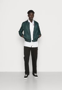 Donkergroene bomberjack, witte button-up shirt, zwarte cargobroek en zwarte sneakers met witte zolen. Soepele texturen en casual ontwerp.