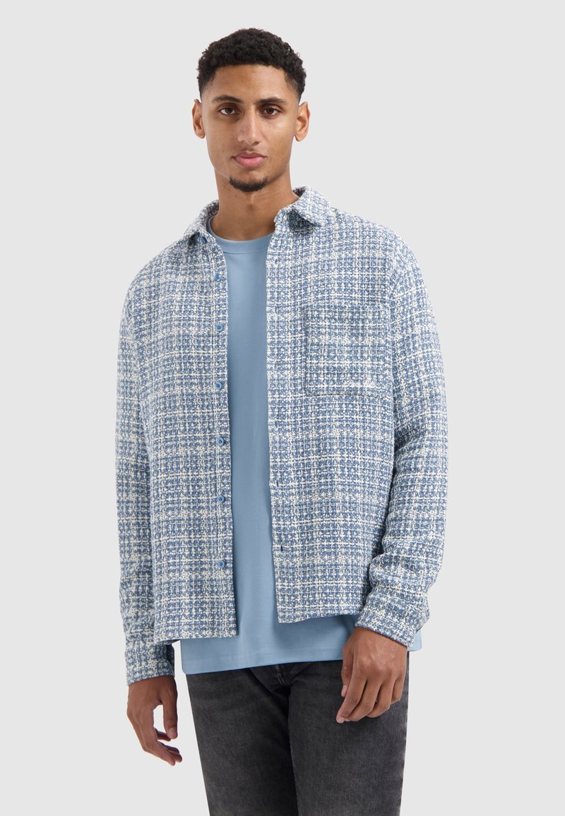 Overhemd met knopen in een blauw-witte gemêleerde stof met een geruit patroon. Bevat een borstzak en heeft een ronde zoom. Draagt over een lichtblauw T-shirt.