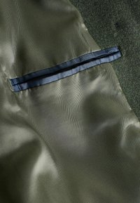 Olivovozelená tkanina s jemnou textúrou, s tmavomodrým saténovým lemom pozdĺž skrytého švíka vrecka.