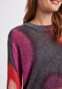 Pull en tricot avec un col rond, fabriqué en tissu doux et duveteux gris, orné de grands motifs circulaires roses et rouges. Texture douce avec une coupe ample.