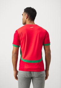 Maglia sportiva rossa con strisce verdi a contrasto. Il tessuto è liscio con un sottile motivo geometrico. Maniche corte e scollatura rotonda.