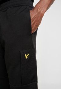 Pantalones de chándal negros con un bolsillo lateral, tejido texturizado y un logo amarillo bordado en la solapa del bolsillo. Cintura elástica visible.