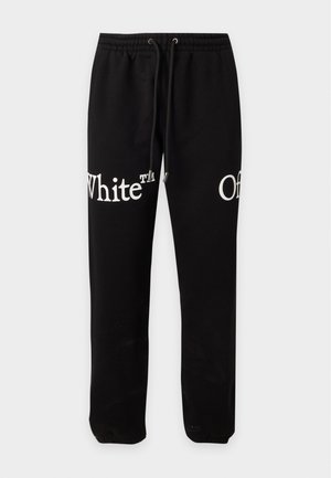 Homme portant un t-shirt blanc Off-White et un pantalon noir avec le texte "Off White", debout, les mains dans les poches sur un fond uni.