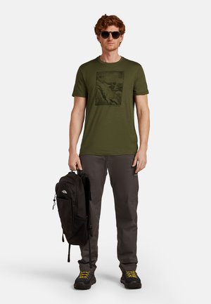 Man met een zonnebril, olijfgroene grafiche t-shirt, donkere broek en wandelschoenen met gele veters, die een zwarte rugzak naast zich houdt.