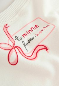 Biały materiał bluzy z prostokątną grafiką, na której znajduje się napis "to: minnie" w kolorze czerwonym oraz "from: santa" w kolorze czarnym, udekorowany czerwonym wzorem wstążki.