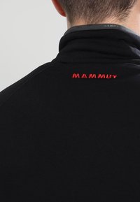 Person som bär en svart jacka med hög krage, med röd "MAMMUT"-logotyp broderad på baksidan nära nacken.