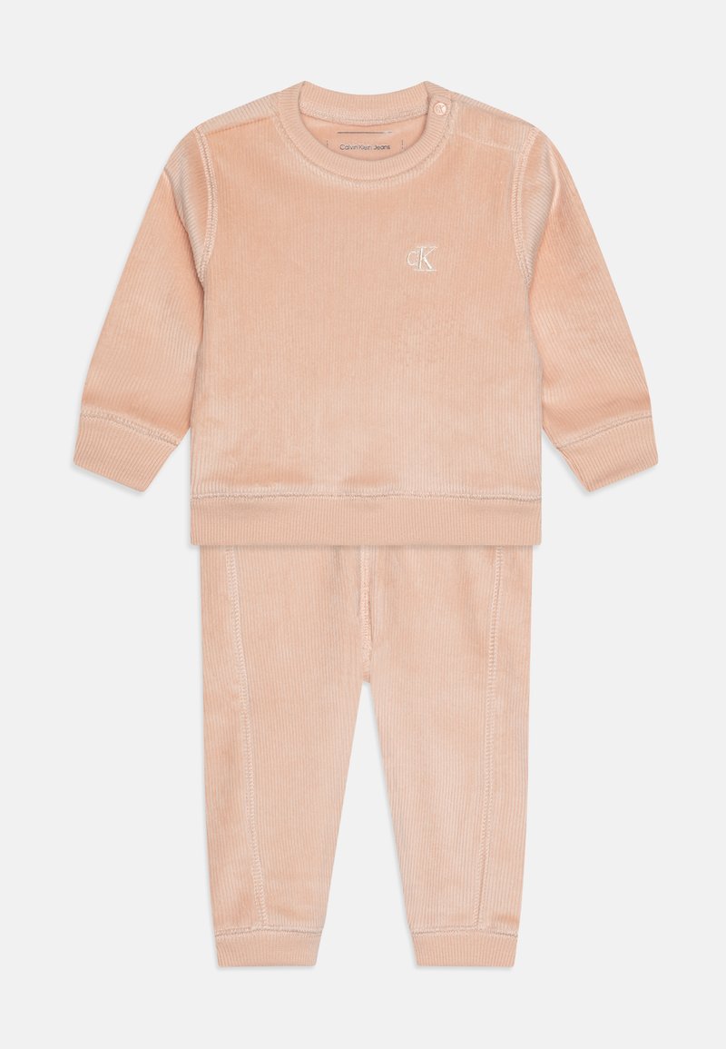 Conjunto de fato de treino em corduroy cor pêssego, apresentando uma blusa de mangas longas com bordas cosidas e calças combinando com punhos elásticos. Gola ajustável.