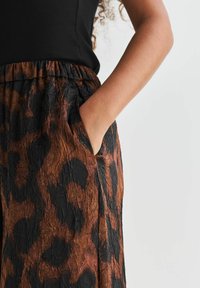 Vrouw in een zwarte top en een gekreukt rokje met een bruin en zwart luipaardpatroon, met haar rechterhand in de zak van het rokje.