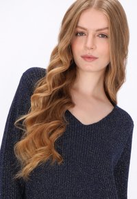 Tengerészkék pulóver bordázott textúrával és V-nyakú dizájnnal. Csillogó szálak finom fényt adnak. Hosszú, puha, hullámos haj részben látható.