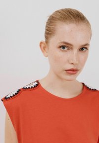 Rød t-shirt med brede ærmer, prydet med blomsterformede hæklede detaljer på skuldrene. Glat tekstur og rund halsudskæring.