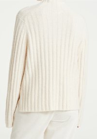 Pull crème en maille côtelée avec un col montant et des épaules tombantes, présentant une texture douce et un ourlet arrière légèrement allongé.