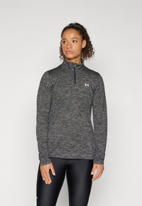 Schwarzes Heideremmershirt mit Viertel-Reißverschluss und langen Ärmeln, glatter Textur, sportlichem Schnitt und Logo auf der Brust, kombiniert mit schwarzen Leggings.