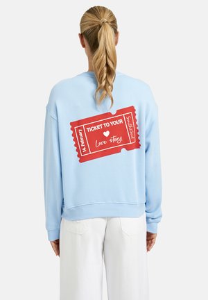 Person with blonde ponytail wearing light blue sweatshirt with red ticket graphic stating "Ticket to your love story 14 February" on back.