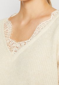 Crèmekleurig V-hals gebreid top met een zachte, textuurafwerking; kantafwerking aan de halslijn voegt detail toe met een verfijnd, geschulpt ontwerp.