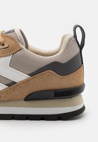 Lähiotoksia beigestä ja ruskeasta lenkkarista, jossa on verkko- ja mokkapaneelit, musta kantalenkki ja paksu kumipohja.