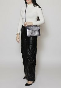 Borsa a tracolla metallica argento con patta, texture liscia e tracolla regolabile, abbinata a un dolcevita bianco aderente e pantaloni in pelle nera.