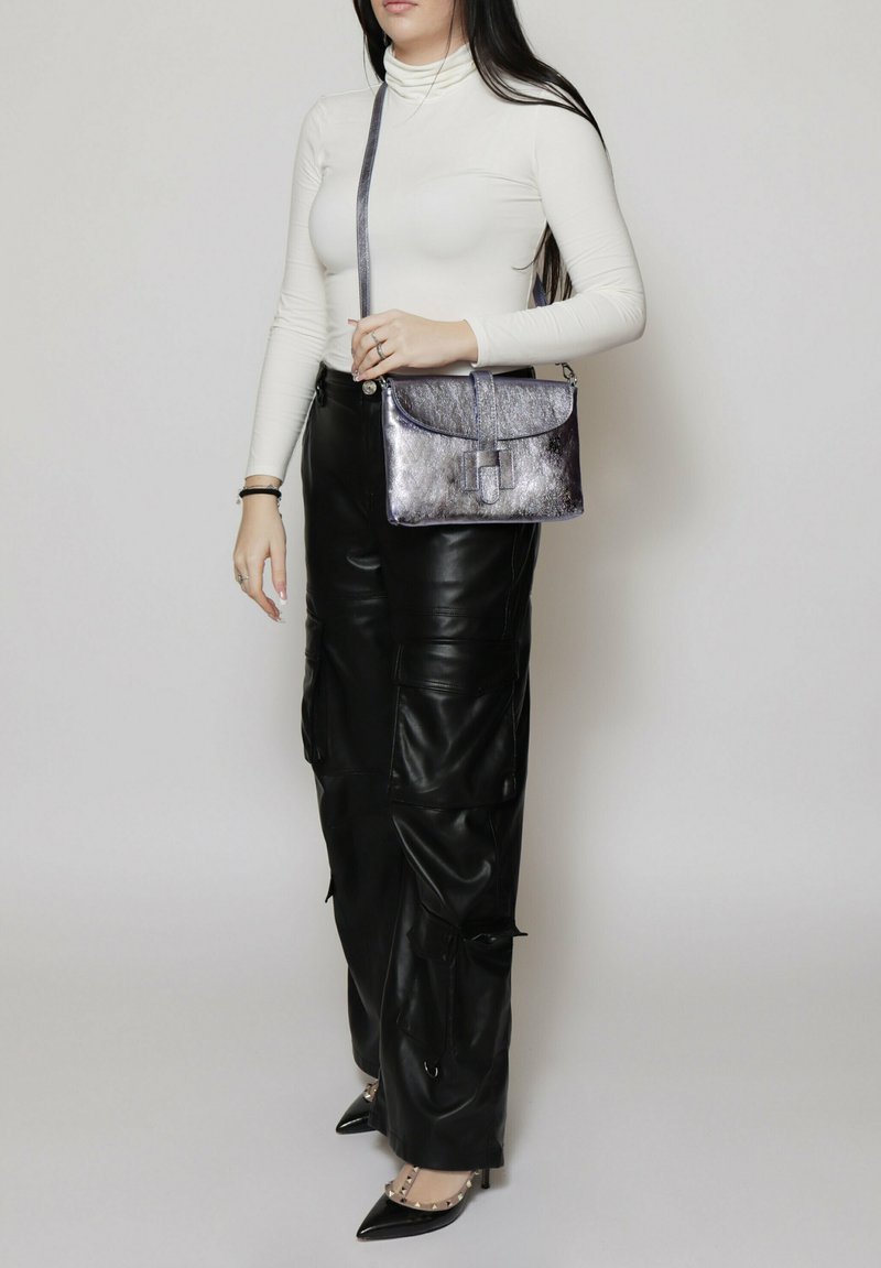 Borsa a tracolla metallica argento con patta, texture liscia e tracolla regolabile, abbinata a un dolcevita bianco aderente e pantaloni in pelle nera.