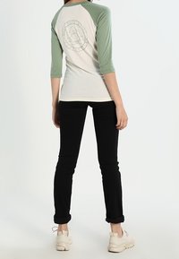 T-shirt raglan a tre quarti, con fronte crema e maniche verdi, presenta un design del logo sul retro. Pantaloni neri aderenti e sneakers chiare.