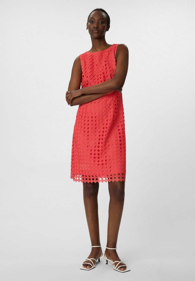 Korallfarbenes Spitzenkleid mit einem diamantenförmigen Ausschnitt-Muster, ärmellosem Design und gewelltem Saum. Kombiniert mit weißen Riemchen-Heels.