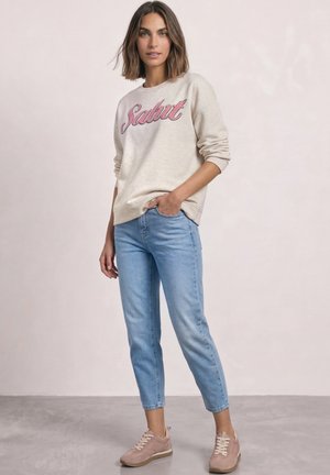 Femme aux cheveux bruns mi-longs portant un sweat-shirt beige avec le texte "Salut", un jean court bleu clair et des baskets roses, debout de manière décontractée.