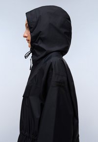 Giacca nera impermeabile con cappuccio, dotata di fondo elasticizzato e un logo sulla manica. Texture liscia e design minimalista.