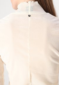 Tessuto color crema con una texture morbida. Presenta un collo arricciato e una chiusura con bottoni sulla parte posteriore. La cucitura corre verticalmente lungo il centro.