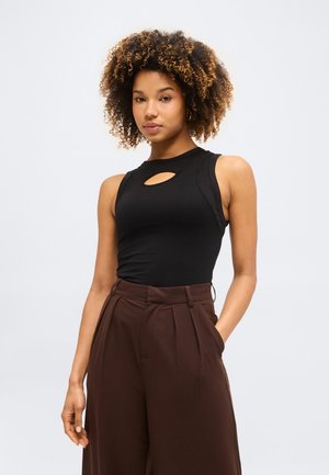 Γυναίκα με σγουρά μαλλιά που φοράει μαύρο αμάνικο τοπ με άνοιγμα στο στήθος και καφέ φαρδιές παντελόνες με ψηλή μέση, στέκεται με το ένα χέρι στην τσέπη.