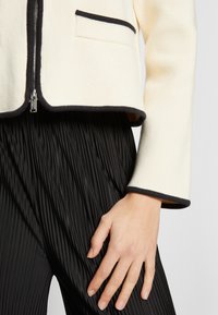 Veste crème avec bordure noire, dotée d'une fermeture éclair et d'une poche ; associée à un pantalon plissé noir, présentant des lignes verticales et une texture lisse.