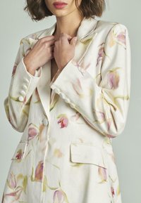 Blazer floral en tejido beige claro, con patrones florales en rosa y verde, dos bolsillos frontales y detalles de botones en las mangas.